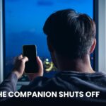 A person alone by a window at night holding a phone with a dark screen, illustrating an AI companion being shut down